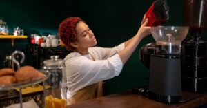 Woman barista filling coffee grinder with beans in a cozy café setting. Internationale Studentin arbeitet als Barista in einem Café
